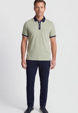 Man draagt een lichtgroen poloshirt met marineblauwe kraag en manchetten, marineblauwe broek en marineblauwe sneakers, staand tegen een effen lichte achtergrond.
