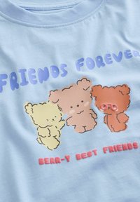 Trois ours dessinés en jaune, marron clair et orange avec des lunettes sous le texte « Amis pour toujours » et « Meilleurs amis comme des ours » sur un tissu bleu clair.