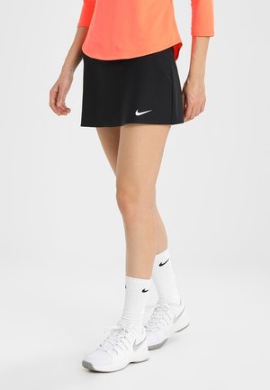 Frau trägt schwarzen Nike Sportsrock, weiße Nike Socken und weiße Nike Sneakers, steht mit einem Fuß leicht nach vorne.