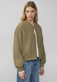 Bomberjack in olijfgroen, lichtgewicht stof, ribgebreide manchetten, ritssluiting, zijzakken, gestyled met een crèmekleurig top en lichtblauwe jeans.