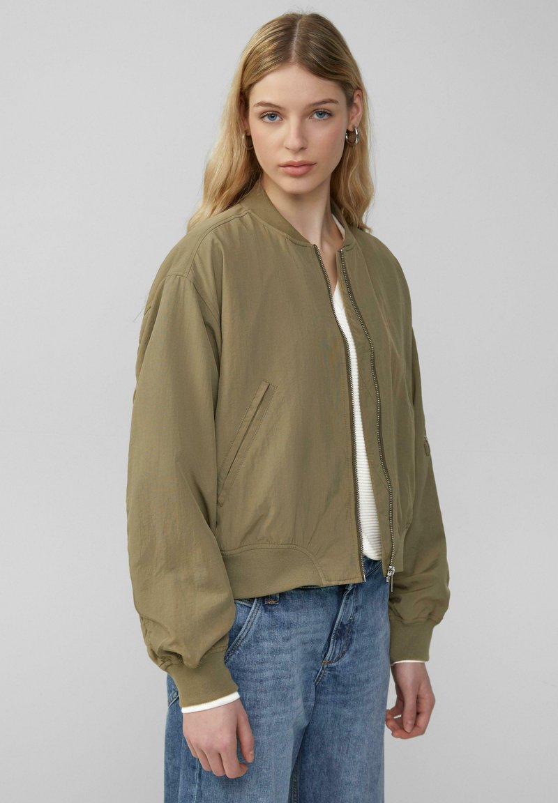 Bomberjack in olijfgroen, lichtgewicht stof, ribgebreide manchetten, ritssluiting, zijzakken, gestyled met een crèmekleurig top en lichtblauwe jeans.