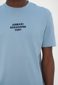 Svjetloplava pamučna majica s okruglim izrezom i kratkim rukavima, s crnim vezom: "ARMANI EXCHANGE 1991" na prednjoj strani.