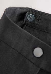 Gros plan de la ceinture d'un pantalon noir montrant un bouton transparent sur une bande élastique réglable et une fermeture par bouton-pression noire.