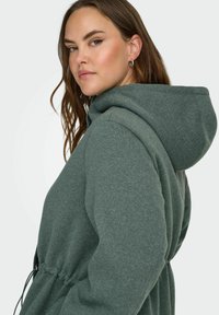 Zeleni kapuča s kordom na pasu, mehkega teksture, dolgimi rokavi in velikim pokrivalom. Ima vidne šive in sproščen kroj.