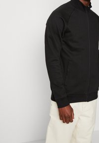 Veste zippée noire avec col montant, poignets côtelés et deux poches latérales ; confectionnée dans un tissu doux et lisse au design minimaliste.