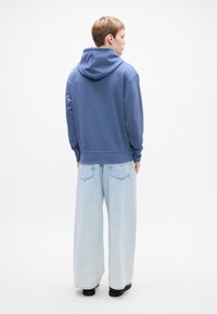 Persona di spalle che indossa una felpa con cappuccio blu, jeans a gamba larga azzurri e scarpe nere su uno sfondo bianco.