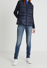 Námořnická péřová vesta s předním zipem, v kombinaci s modrými úzkými džínami a bílými teniskami, na neutrálním pozadí.