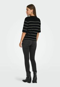Woman standing in black skinny jeans, black boots, and a black short-sleeve top with white horizontal stripes, facing sideways with head turned.