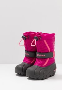 Botas de invierno aisladas en color rosa con suelas de goma negra, que cuentan con un borde ajustable con cordón, tiras de Velcro etiquetadas "SOREL" y suelas con textura para un mejor agarre.