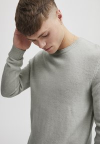 Pull en tricot gris clair, avec un col rond et des poignets côtelés, présentant une texture douce et une coupe ajustée.
