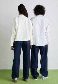 To figurene står rygg mot rygg. Den ene har på seg en kremfarget bomullsjakke og mørkeblå jeans. Den andre har en lyst stripete skjorte og denimbukser.