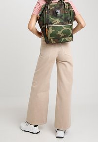 Henkilö, jolla on päällä vaaleanbeiget leveälahkeiset housut, valkoiset lenkkarit, vaaleanpunainen lyhythihainen paita ja vihreä maastokuvioinen reppu, seisoo kädet lanteilla.