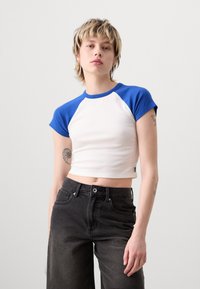 Maglietta bianca a maniche corte con maniche raglan blu, caratterizzata da un design aderente e una morbida texture. Indossata con jeans neri in denim a vita alta.