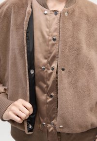 Smeđa bomber jakna s mekim, krznastim vanjskim slojem i glatkom unutarnjom postavom; sadrži gumbe s kopčanjem i rebraste manžete.