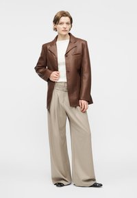 Γυναίκα με καφέ δερμάτινο σακάκι, λευκό υφασμένο τοπ, μπεζ παντελόνι φαρδιάς γραμμής και μαύρα παπούτσια, που στέκεται μπροστά σε απλό ανοιχτό φόντο.