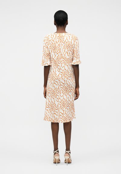 Γυναίκα όρθια με την πλάτη στην κάμερα, φορώντας λευκό φόρεμα μεσαίου μήκους με πορτοκαλί πουά και μπεζ πέδιλα με τακούνια.