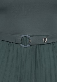 Jupe plissée vert foncé avec ceinture en tissu assortie fixée par un anneau rond foncé et une fermeture à bouton-pression métallique à la taille.