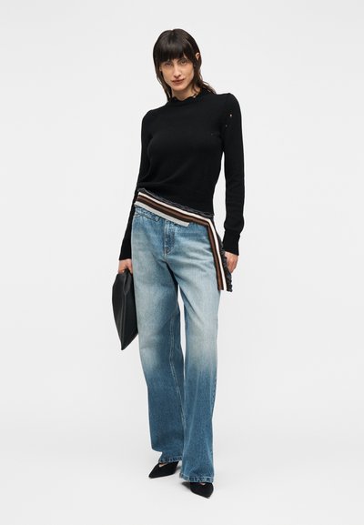 Pulover negru tricotat cu decolteu ondulat, asortat cu blugi largi de culoare albastru deschis, prevăzuți cu o curea cu dungi. Ține o geantă tip clutch neagră.