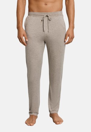 Mężczyzna w jasnoszarych spodniach dresowych ze ściągaczem, stojący boso na jednolitym jasnym tle, widoczny od połowy torsu do stóp.