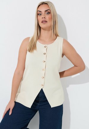 Lyshåret kvinde iført ærmeløs cremefarvet bluse med knapper og mørkeblå jeans, poserer med den ene hånd på hoften mod en hvid baggrund.