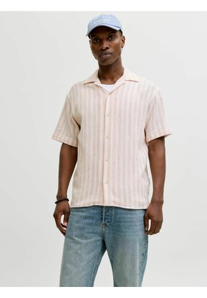 Mężczyzna w jasnej, paskowanej, krótkorękawowej koszuli na guziki, niebieskich dżinsach, jasnoniebieskiej czapce i czarnej bransoletce na tle jednolitego tła.