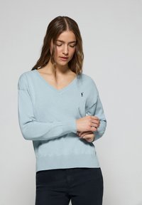 Pull-over léger bleu clair en V avec des manches longues, des poignets et un ourlet côtelés. Présente un petit logo noir sur la poitrine gauche. Texture douce.