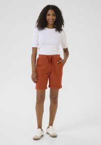 Shorts couleur rouille avec une taille à cordon, poignets retroussés et poches avant, associés à un haut à manches courtes en ribiscage blanc et des baskets décontractées.
