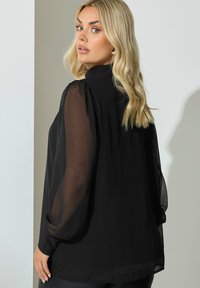 Blusa negra con mangas abullonadas transparentes y un cuello alto. Presenta un corte holgado y puños con botones. Textura suave con una silueta fluida.