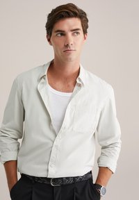 Šviesiai pilka marškinėlių viršutinė dalis su krūtinės kišeniu, balta marškinėlių apatinė dalis, juodas pintas diržas ir juodos kelnės. Lygus audinys su atsitiktiniu siluetu.