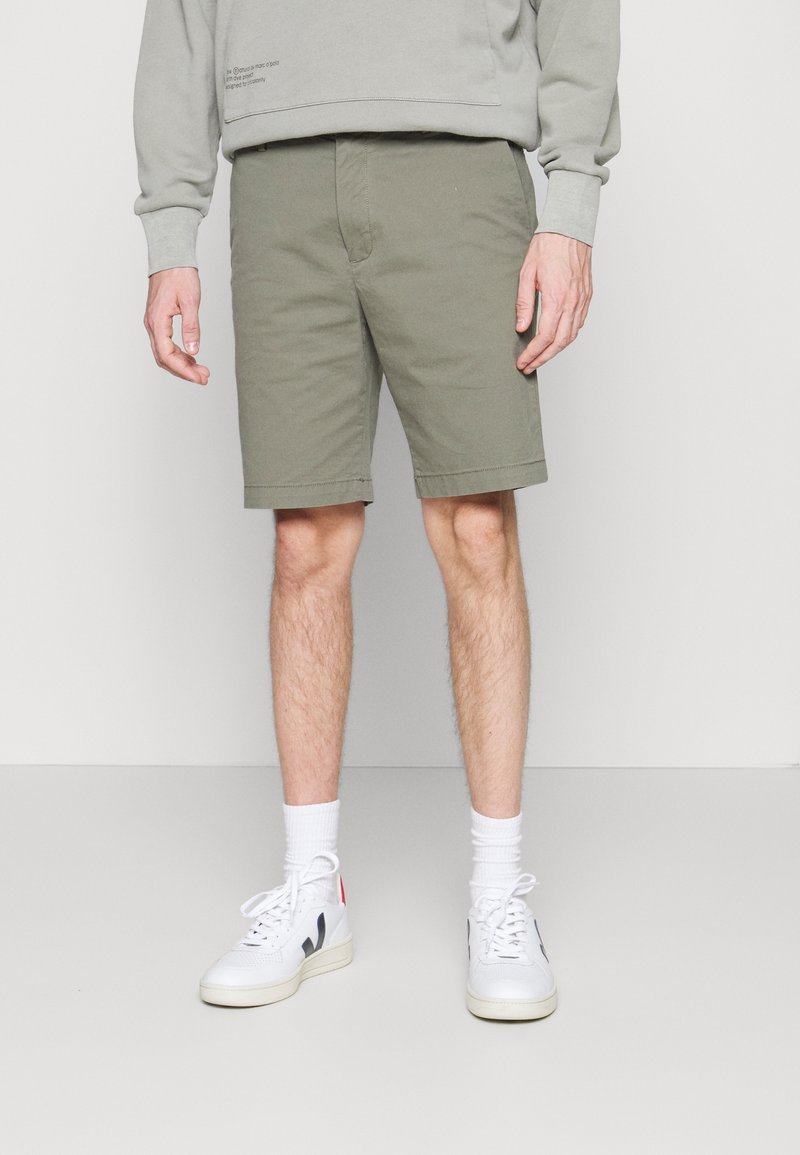 Homme portant un short vert olive, des chaussettes hautes blanches et des baskets blanches avec des détails noirs et rouges, debout devant un fond uni.