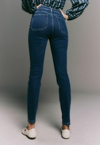 Džínové kalhoty v tmavě modré barvě s vysokým pasem, úzkým střihem a dvěma zadními kapsami. Kombinované s bílými botami, vystavené na neutrálním pozadí.