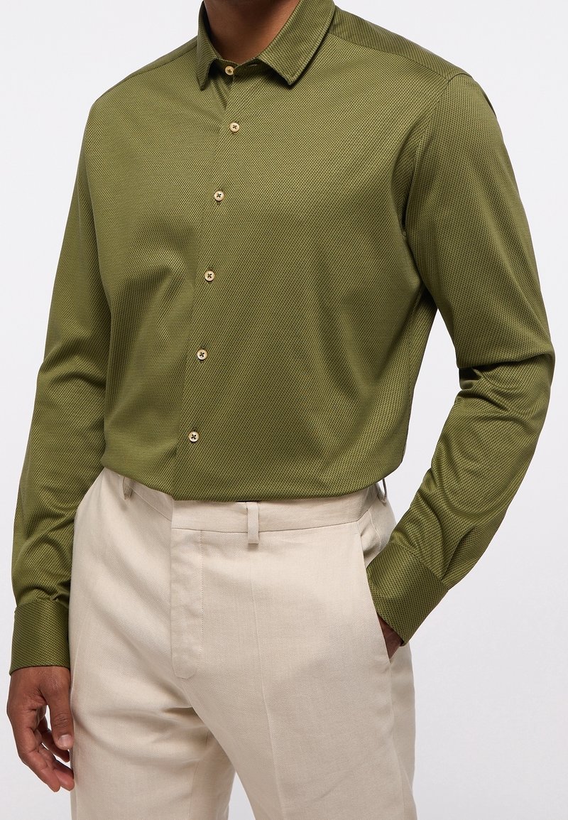 Camicia verde oliva con bottoni in tessuto testurizzato, caratterizzata da un colletto e bottoni in tonalità legno, abbinata a pantaloni leggeri beige.