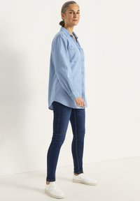 Ljusblå denimskjorta med knappar, långa ärmar och två bröstfickor, matchad med mörkblå slim-fit jeans och vita sneakers.