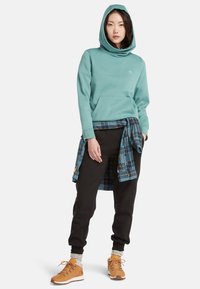Un sweat à capuche turquoise avec un col large, une poche avant et un pantalon de survêtement noir. Superposé avec une chemise à carreaux bleue nouée à la taille et des bottes de randonnée beiges.