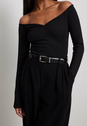 Kvinde iført en sort off-shoulder langærmet top, der er stoppet ned i højtaljede sorte bukser med et sort læderbælte og guldlås.