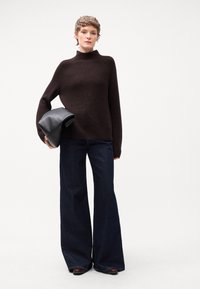 Maglione a collo alto marrone a costine, vestibilità ampia, abbinato a pantaloni a zampa d'elefante in denim scuro. Tiene una pochette nera in pelle. Stile semplice e moderno.