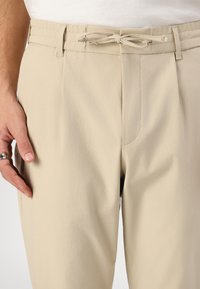 Beige kalhoty s elastickým páskem na šňůrku, střižené do pasu, hladký povrch, boční kapsy a decentní šití na přední straně.