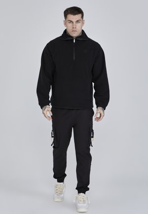 Svart fleecejacka med hög krage och halvmudden, tillsammans med svarta cargobyxor med sidofickor och vita sneakers.
