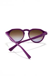 Violetiniai plastikinių saulės akinių suapvalintais geometriniais rėmeliais ir gradiento rudais lęšiais. Šventyklos plonos ir pažymėtos užrašu „HAWKERS“.