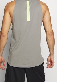 Muž v šedém bezrukávovém tričku Under Armour a černých sportovních šortkách, zobrazený zezadu na jednobarevném pozadí.