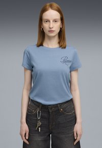 Lichtblauw T-shirt met korte mouwen met "Puma" geborduurd in donkere draad. Gecombineerd met donkere spijkerbroek. Sleutelhanger bevestigd aan de riemlus.