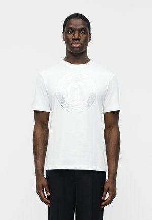 Άνδρας που φοράει απλό λευκό κοντομάνικο μπλουζάκι με διακριτικό ανάγλυφο κυκλικό λογότυπο και μαύρο παντελόνι, στέκεται μπροστά σε λευκό φόντο.