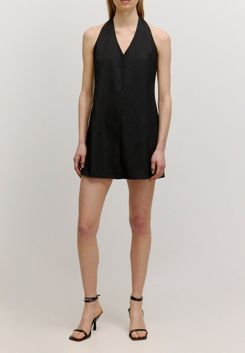 Mini-robe noire à dos nu avec une texture raffinée et un profond décolleté en V, dotée d'une coupe droite et de lignes épurées. Talons noirs à brides.