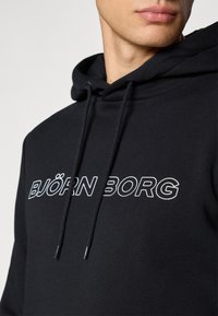 Gros plan sur une personne portant un hoodie noir avec le texte blanc "BJÖRN BORG" et des cordons visibles autour de la zone du cou.