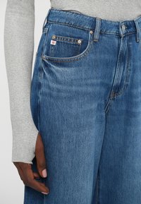 Jeans din denim albastru cu picioare largi și un finisaj decolorat, dotate cu nituri din cupru, un tab de logo mic și o textură subtilă.