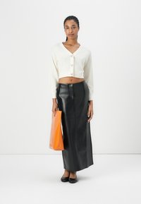 Cardigan courte tricoté blanc avec des manches bouffantes, associé à une longue jupe en cuir noire et un sac fourre-tout orange. Des ballerines noires classiques complètent le look.