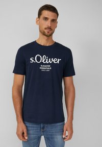 Navyblauwe katoenen T-shirt met een witte s.Oliver-logo en de tekst "ELEVATED ESSENTIALS SINCE 1969." Klassieke ronde halslijn en korte mouwen.