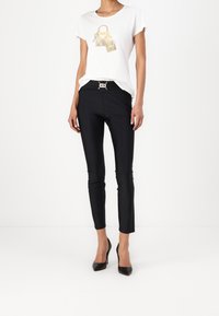 Žena v bílém tričku s potiskem zlatých kabelek, černých slim-fit kalhotách s předním stahováním a černých vysokých podpatkách.