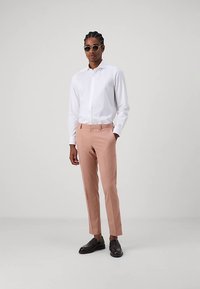 Weißes Hemd mit langen Ärmeln, hellrosa maßgeschneiderte Hose und schwarze Anzugschuhe. Mit runden Sonnenbrillen accessoriert.