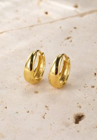 Boucles d'oreilles en or en forme de créole avec une finition lisse et polie, présentant un design courbé et un fermoir sécurisé. La surface présente une texture réfléchissante.